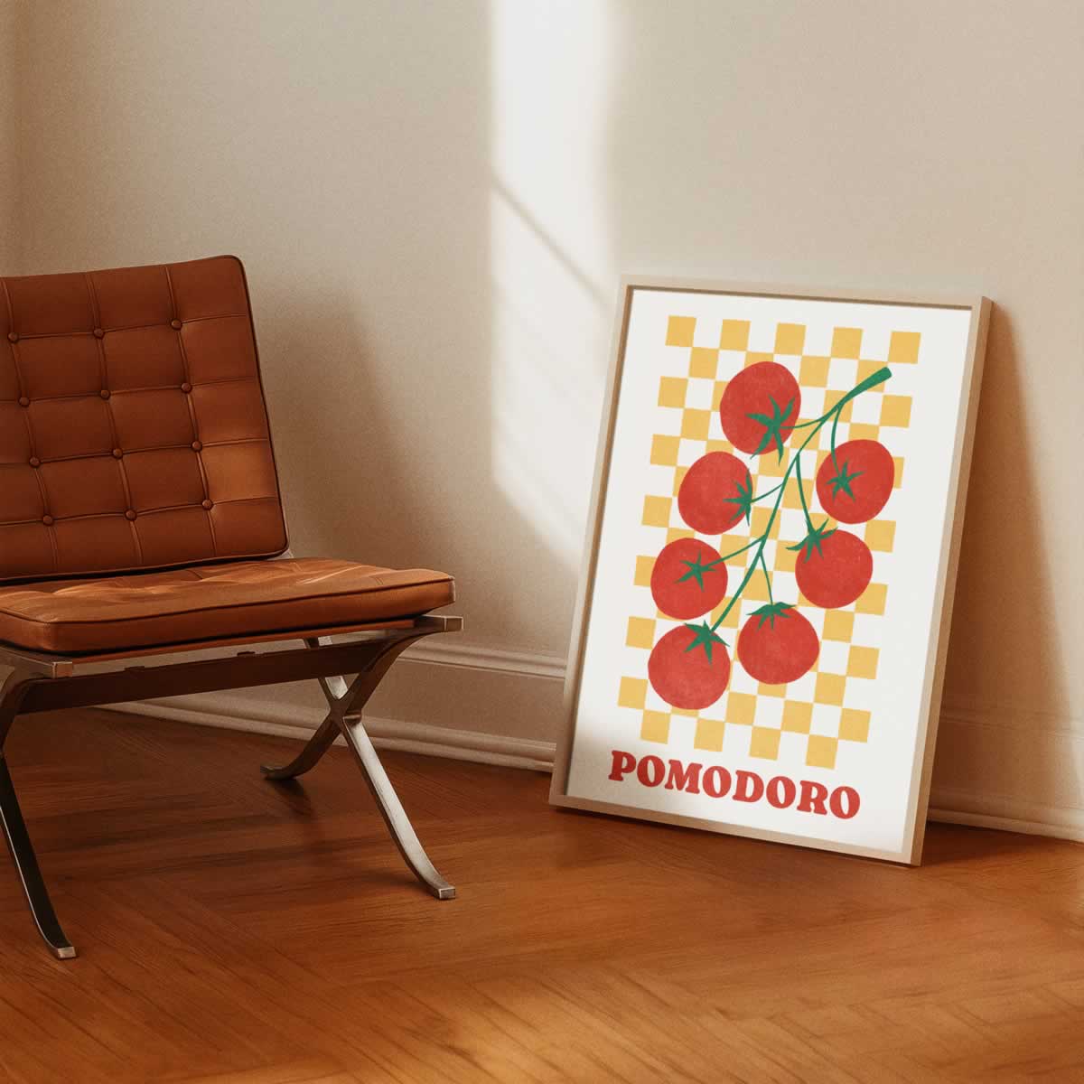 A framed art print featuring a stylised illustration of red tomatoes on a yellow checkerboard background with the word 'POMODORO' in red, leaning against a beige wall on a wooden floor next to a brown leather chair.