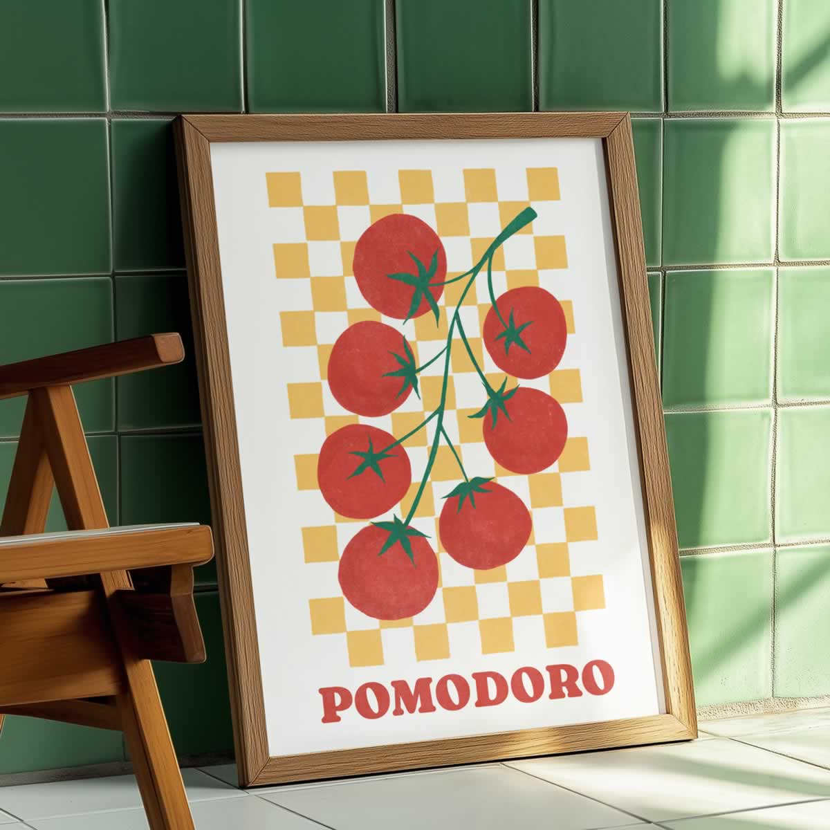 A framed art print featuring a retro-style illustration of red tomatoes on a yellow chequerboard background with the word 'POMODORO' in red at the bottom, leaning against a green tiled wall next to a wooden chair.