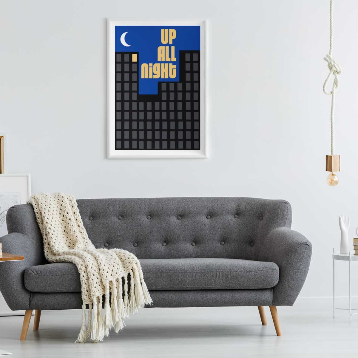 A framed minimalist art print with the text 'UP ALL night' in yellow on a blue and black cityscape background, hanging on a white wall above a gray tufted sofa with a cream knit throw blanket.